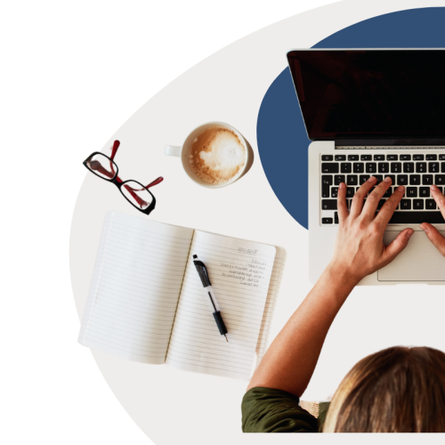 Person sitzt an Laptop, neben ihr liegt eine Brille, Notizbuch, Kaffetasse